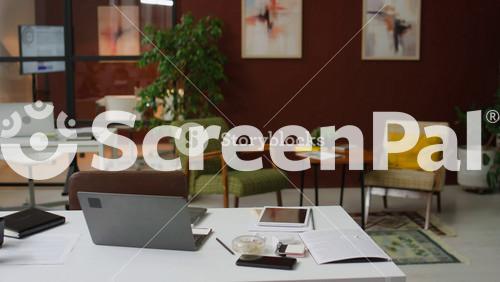No People View Of Interior Of Modern Office With White Work Desk With Laptop, Notebook, Digital Tablet, Phone And Business Documents On It, Another Table, Two Armchairs And Coffee Table In Background